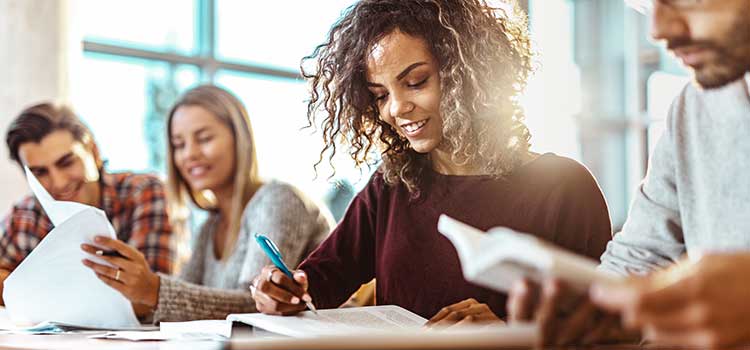 female student writing in notebook at table with three other students