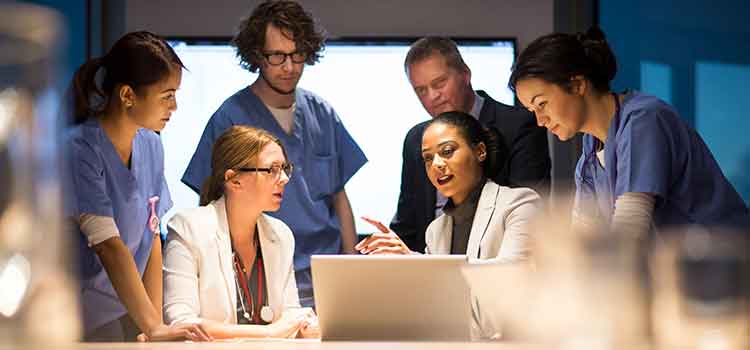 two female medical workers in discussion over laptop with other staffers standing behind them 